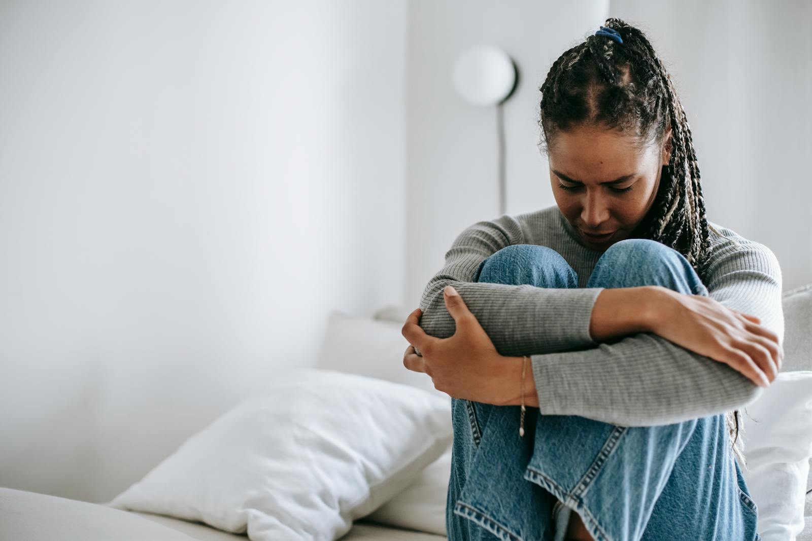 A young woman embraces her knees while sitting on a bed, appearing thoughtful and introspective.