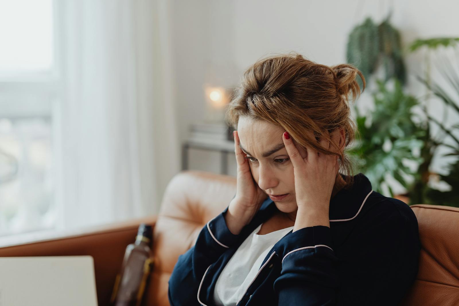 A woman in pajamas sits indoors with hands on head, showing stress and frustration. Perfect for mental health themes.