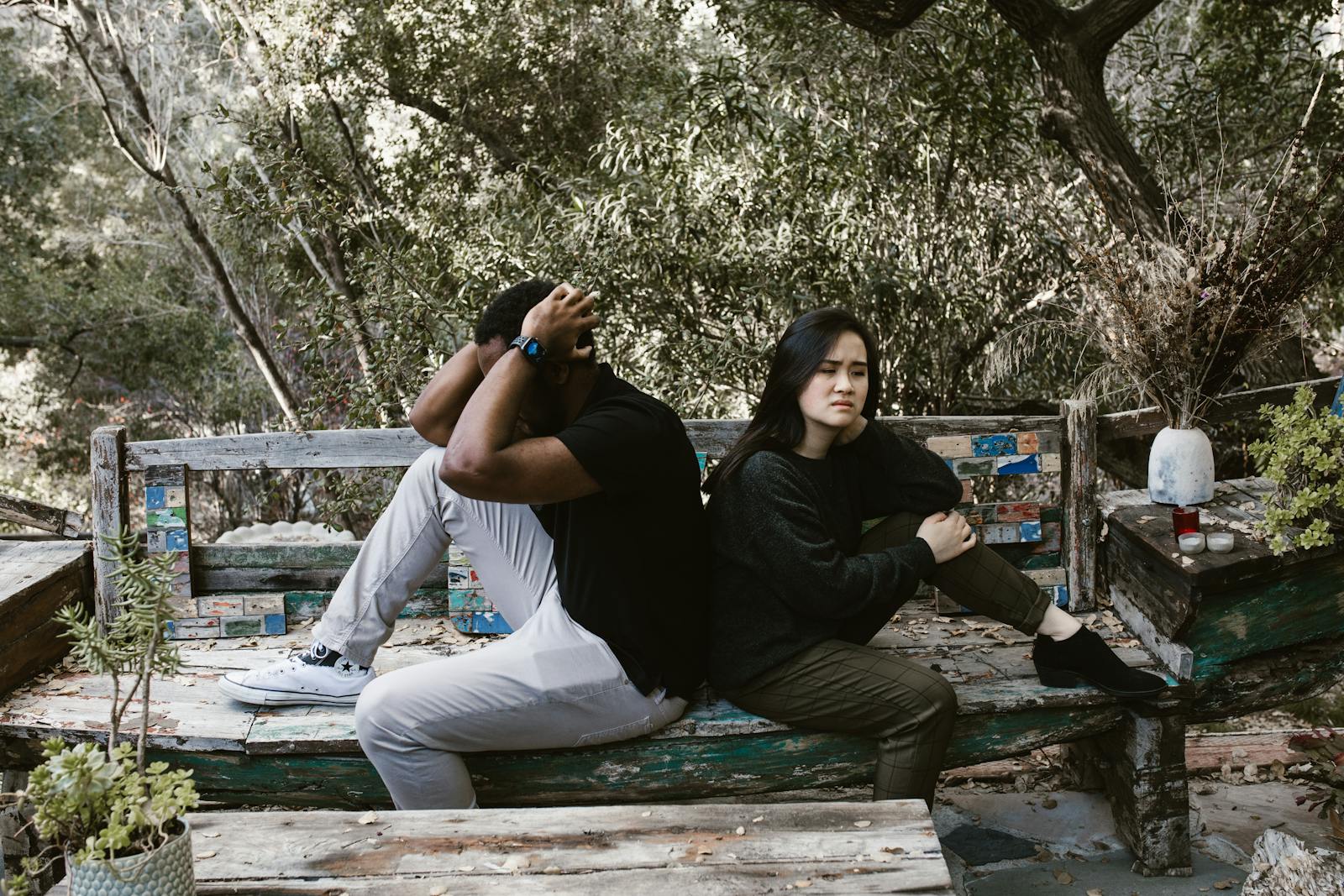 A diverse couple sits on a wooden bench outdoors, expressing anger and sadness.