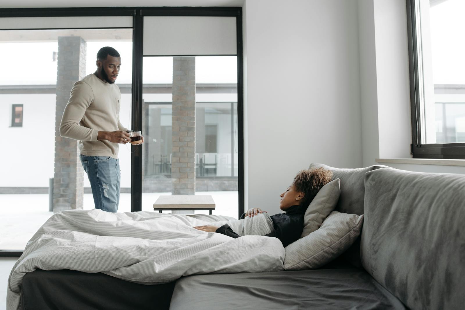 A couple in a modern home setting with one person caring for their sick partner on the sofa.