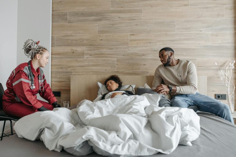 A healthcare worker caring for a sick person in bed, with a supportive partner nearby.