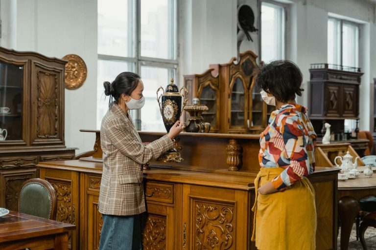 Two women wearing masks browse an antique store, examining a decorative vase.