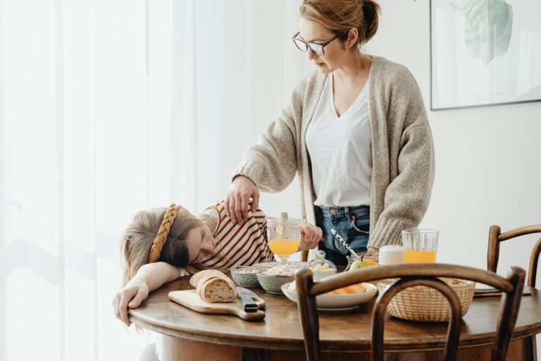 A mother gently wakes her sleepy daughter at a breakfast table filled with healthy foods.