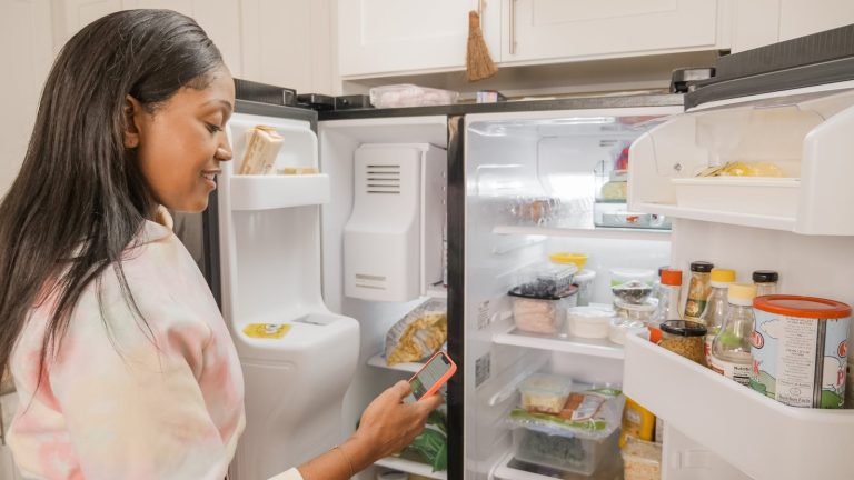 A woman stands at the open fridge in a kitchen, checking her phone.