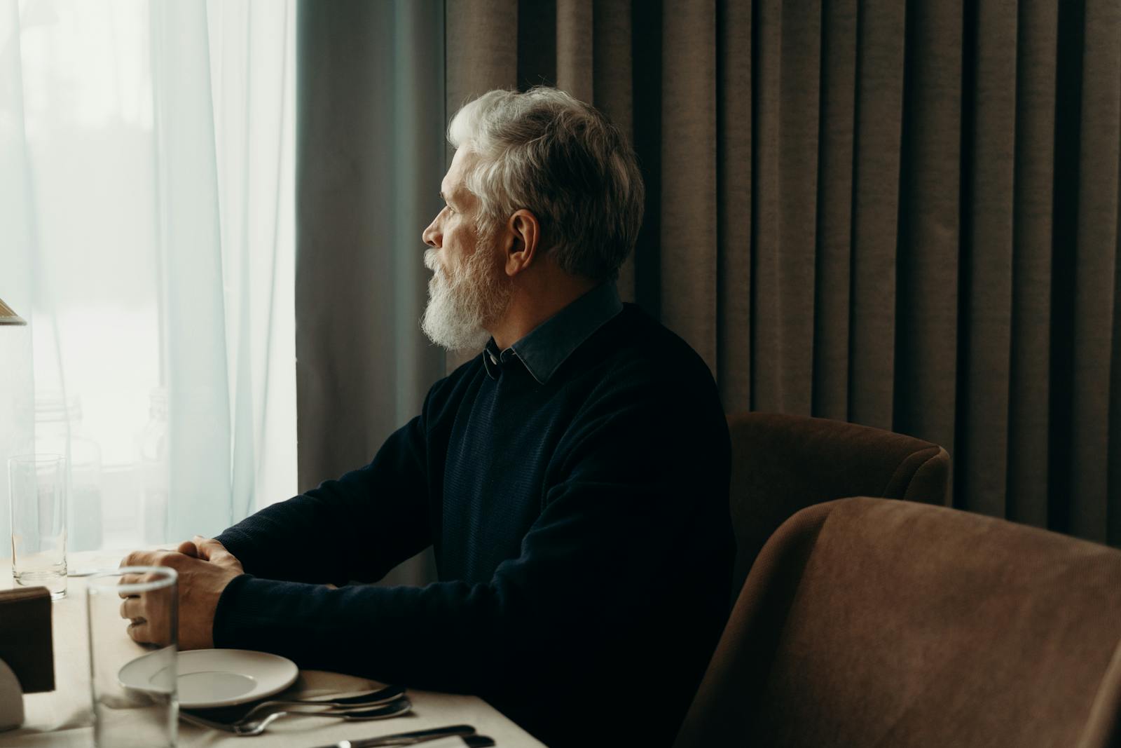 An elderly man with a beard looking outside while sitting by a table indoors.