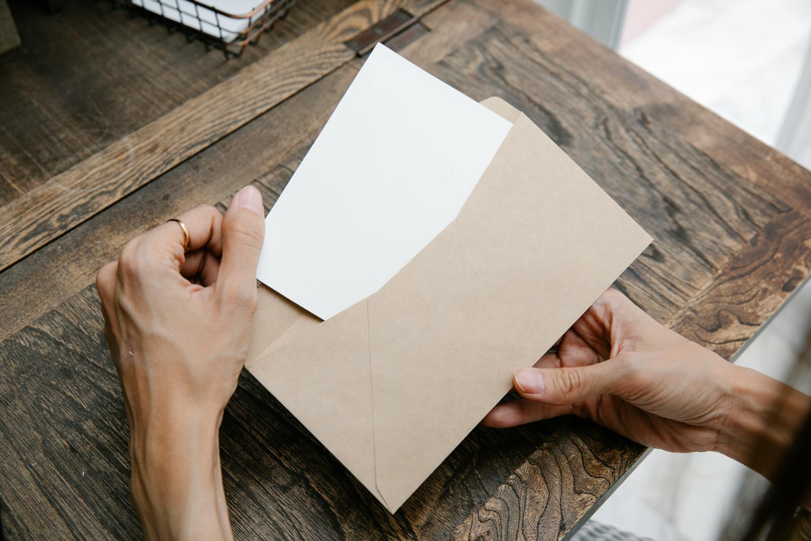 Hands holding a letter inside a brown envelope on a wooden desk, viewed from above.