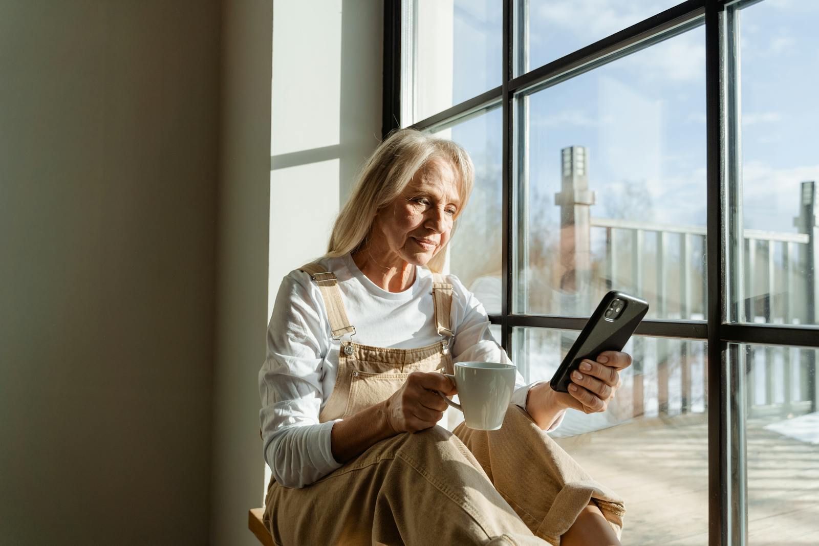 Elderly woman relaxing with coffee and smartphone by a sunny window.