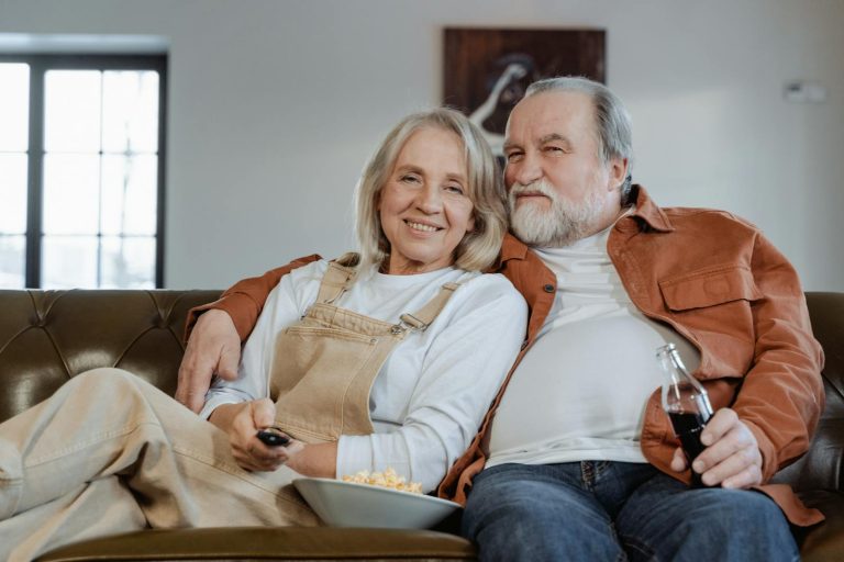 Happy elderly couple enjoying a cozy evening at home, watching TV with popcorn and drinks.