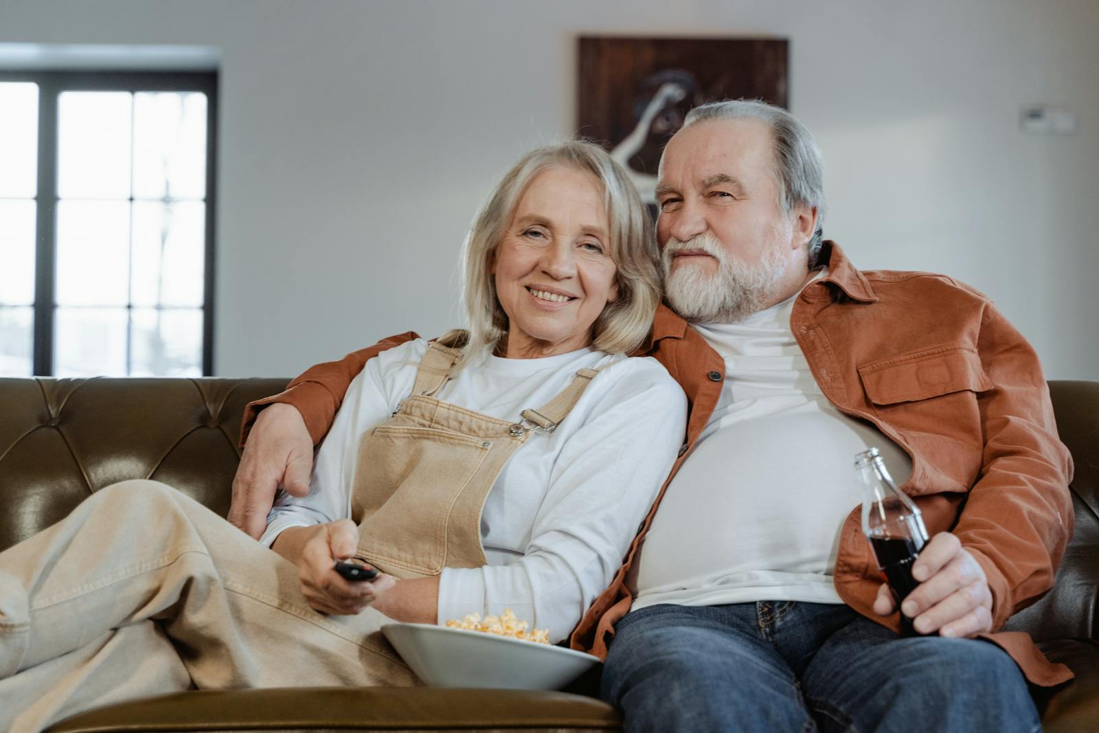 Happy elderly couple enjoying a cozy evening at home, watching TV with popcorn and drinks.