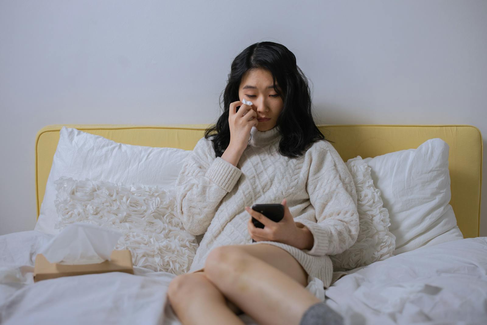Asian woman in a sweater, looking sad and wiping tears, holding a smartphone seated on a bed.