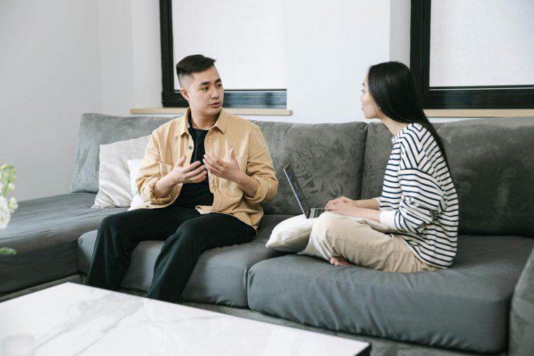 Two young adults engaged in a conversation, sitting on a couch at home.