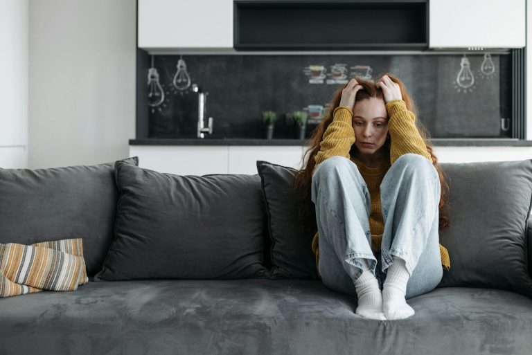 A woman in a yellow sweater sits on a couch, looking sad and pensive, indoors.