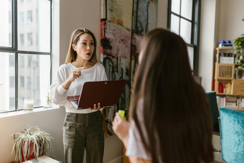 Frustrated mother working on a laptop while talking to her daughter indoors.