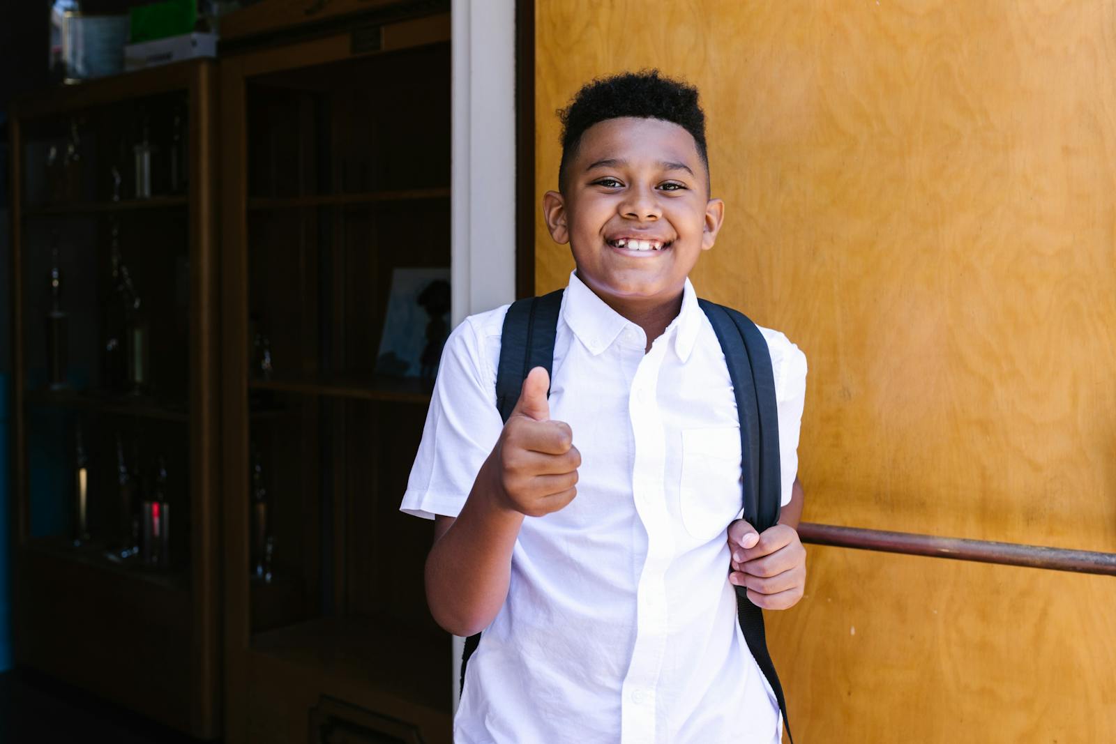 Smiling student with backpack giving thumbs up, standing at school entrance.