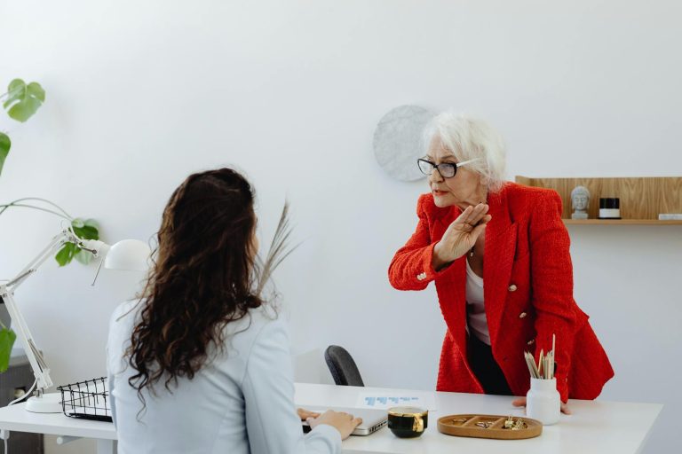An elderly woman in a red jacket expressing displeasure in an office setting with a colleague.