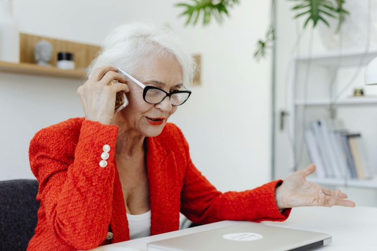 Elderly woman in red blazer talking on phone, working at desk with laptop in office.