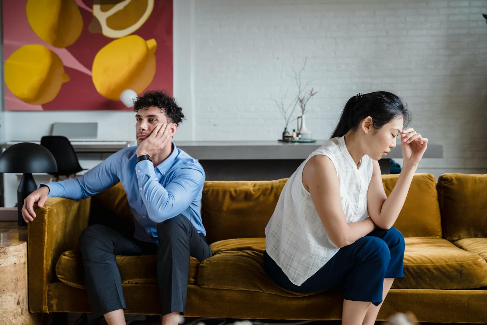 A couple sits apart on a sofa, looking unhappy and deep in thought, reflecting relationship conflict.