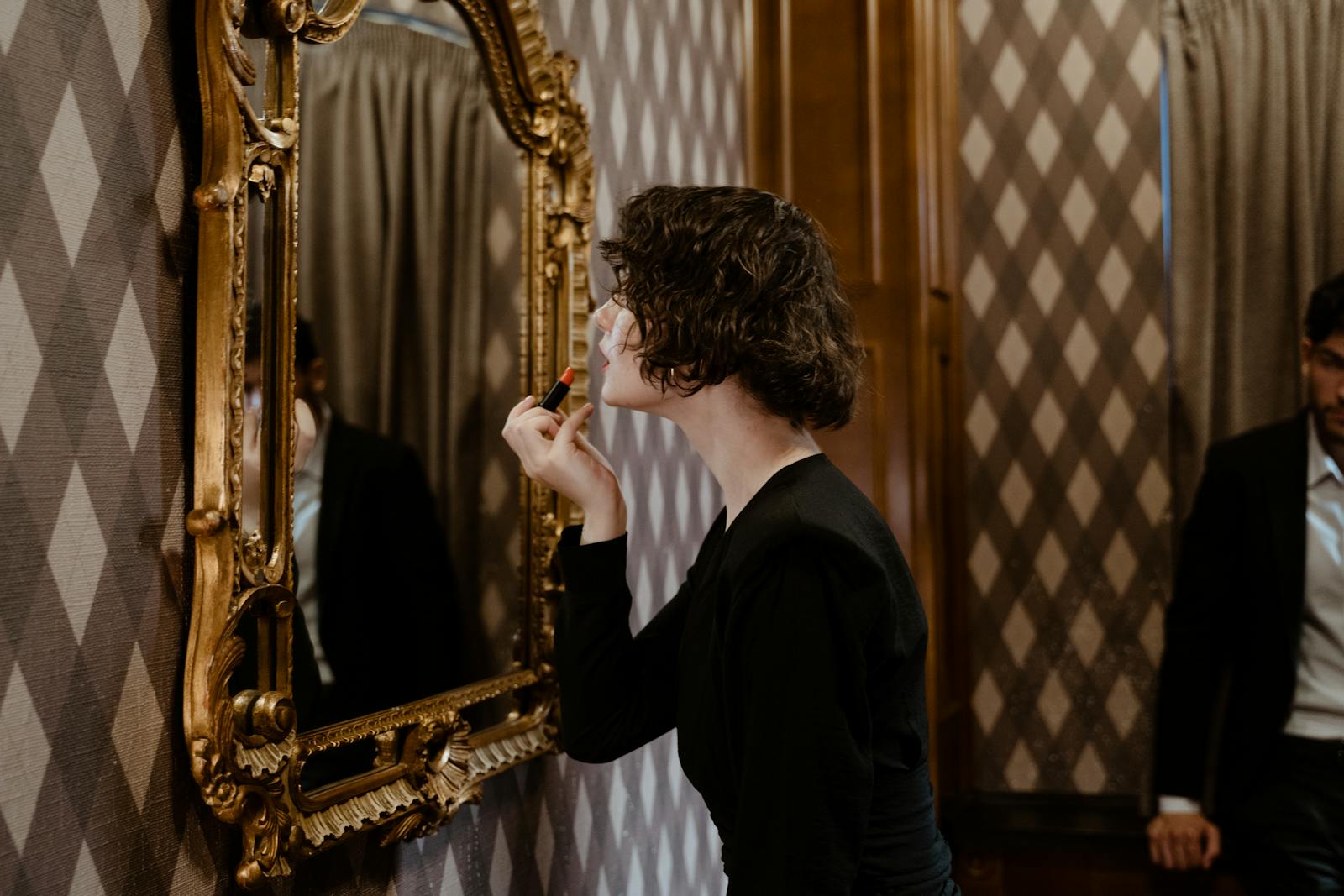 A woman in a black dress applies lipstick in a vintage-style mirror, highlighting elegance and refinement.
