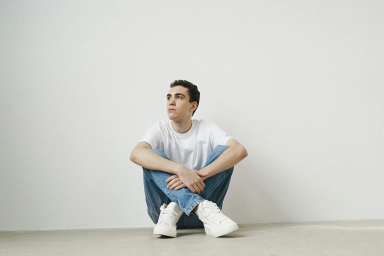 Young man posing casually in a white shirt and jeans against a minimalist white background.