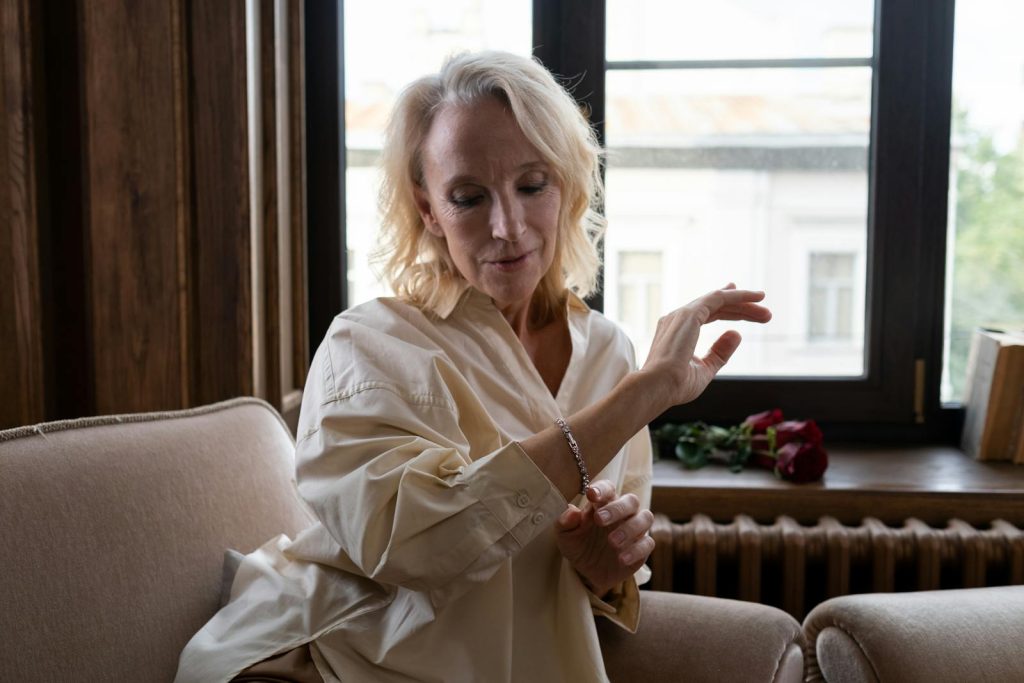 A senior woman sitting indoors, admiring a jewelry bracelet with roses nearby.