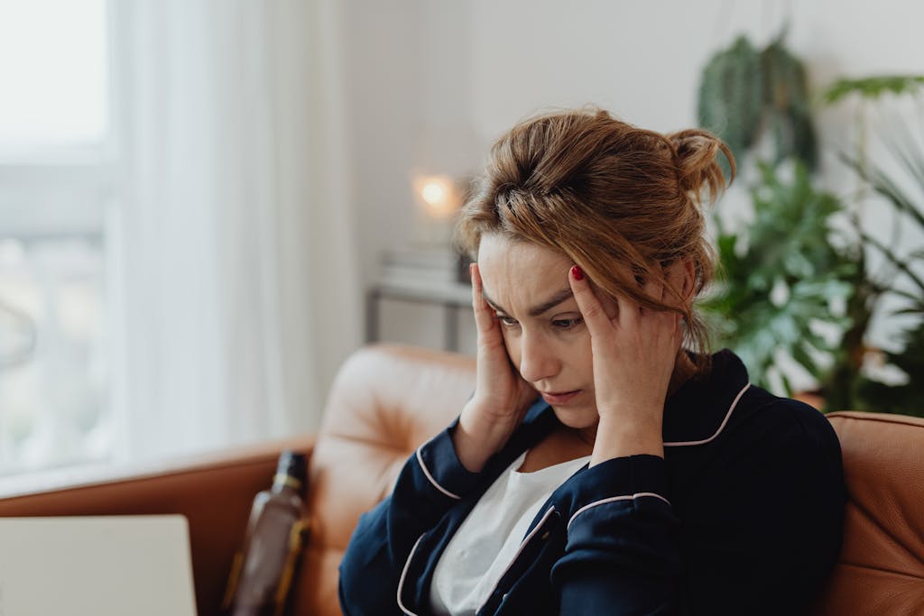 A woman in pajamas sits indoors with hands on head, showing stress and frustration. Perfect for mental health themes.