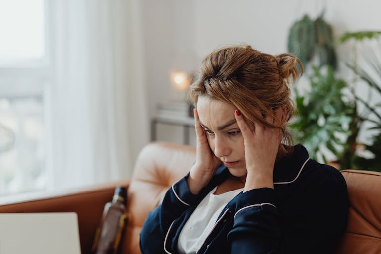 A woman in pajamas sits indoors with hands on head, showing stress and frustration. Perfect for mental health themes.