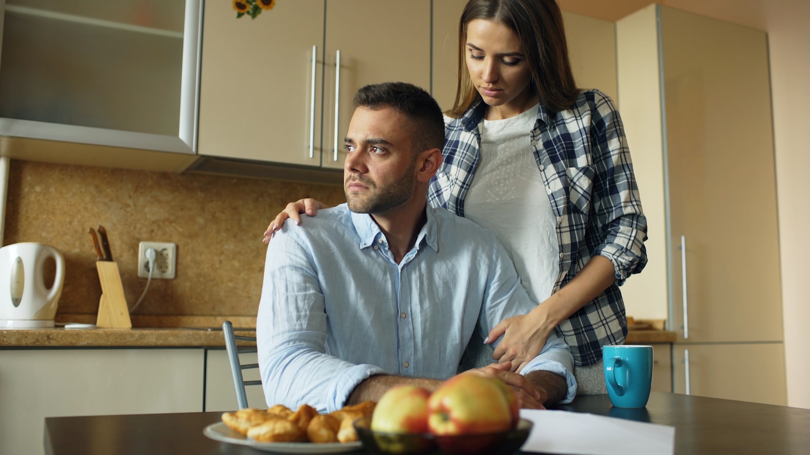 Woman comforts man at kitchen table with food.