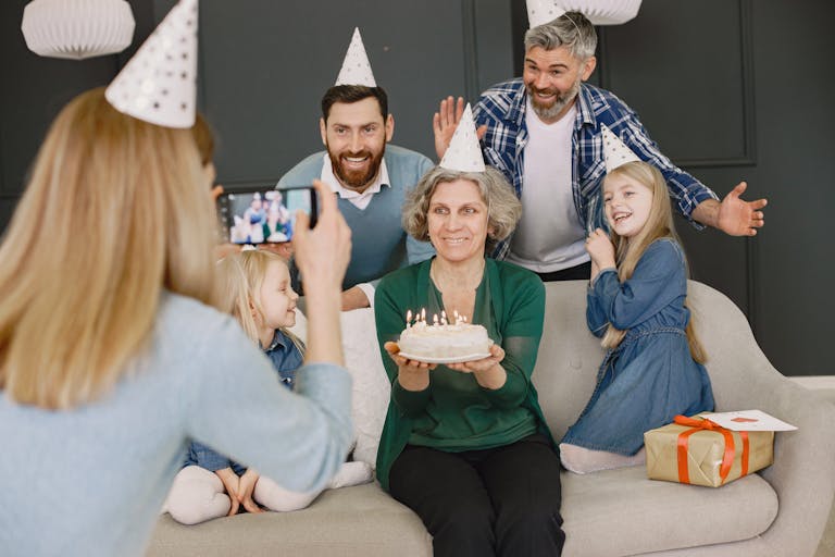 Grandmother holding cake, surrounded by family in party hats, smiling for a photo.