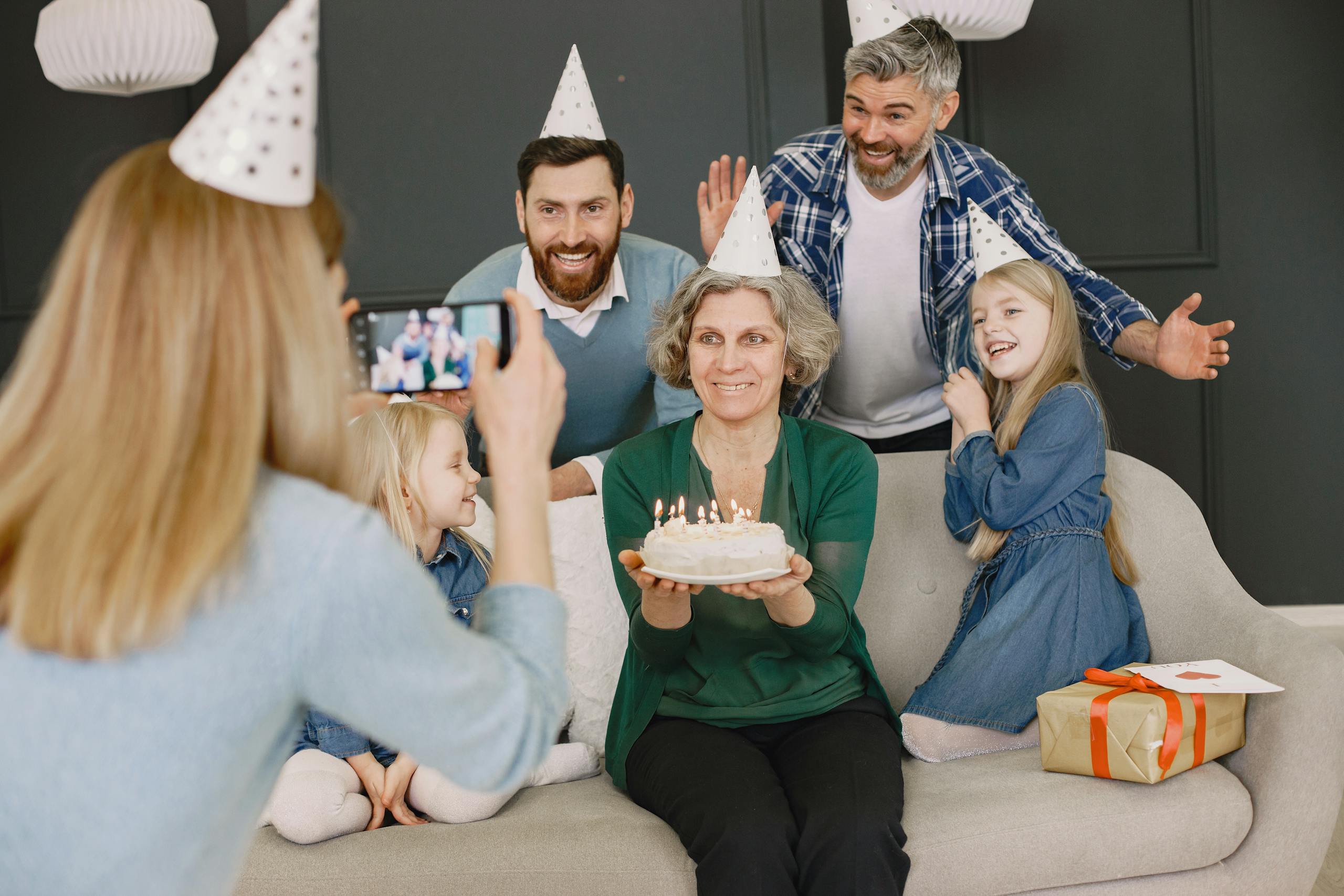 Grandmother holding cake, surrounded by family in party hats, smiling for a photo.