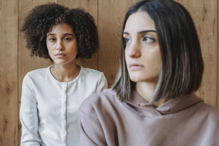 Two young women sit pensively indoors against a wooden wall, deep in thought.