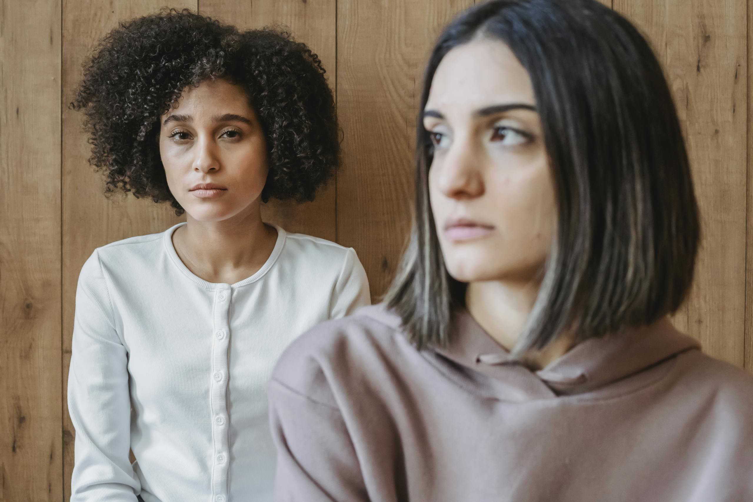 Two young women sit pensively indoors against a wooden wall, deep in thought.