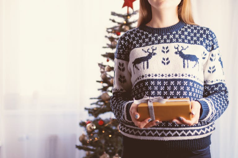 Woman holding a wrapped present in front of a decorated Christmas tree indoors, wearing a festive sweater.