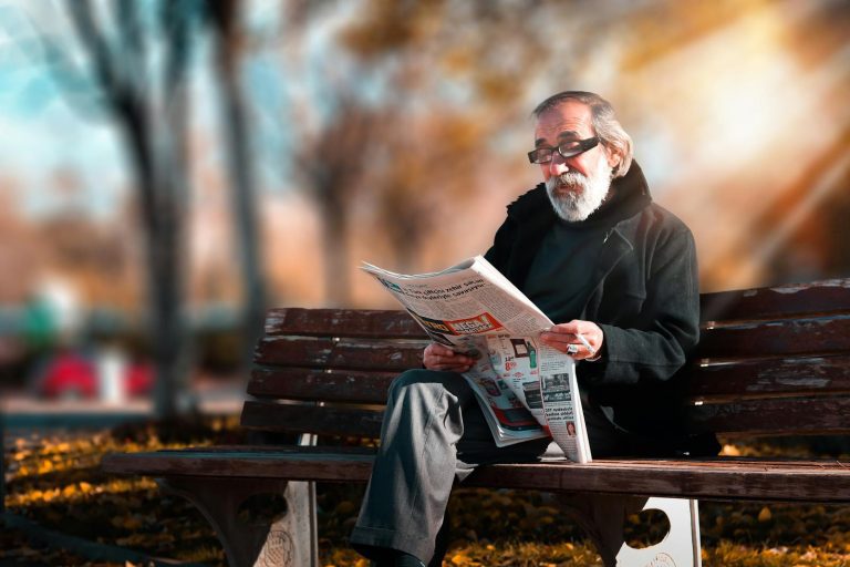 Senior man with beard reading a newspaper on a wooden bench in a sunny park setting.