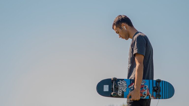 Teenager holding a skateboard, wearing AirPods, under clear blue sky.