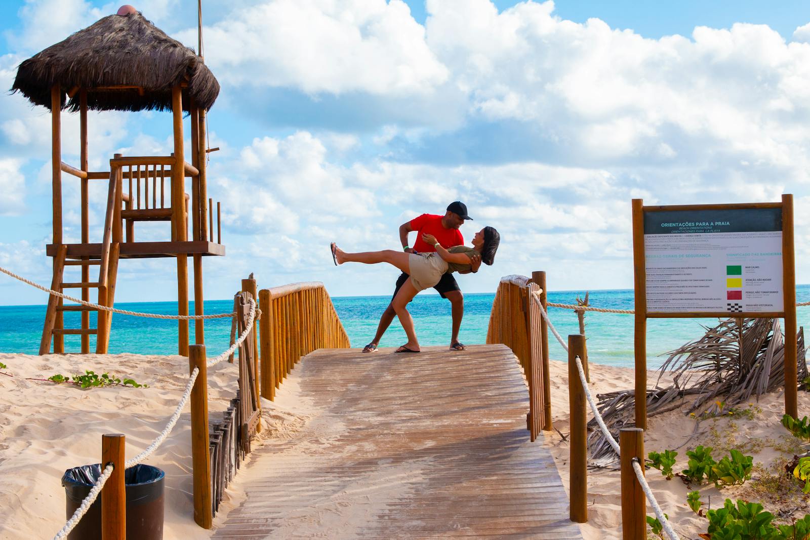 Couple embracing on a wooden bridge at a beach in Natal, Brazil, with vibrant turquoise water and a lifeguard stand.