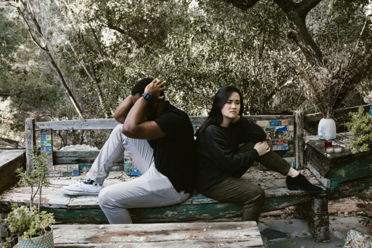A diverse couple sits on a wooden bench outdoors, expressing anger and sadness.