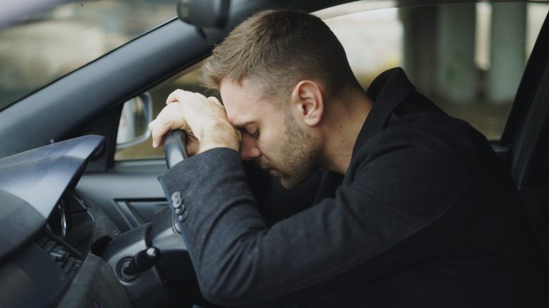 Man resting head on steering wheel in car.