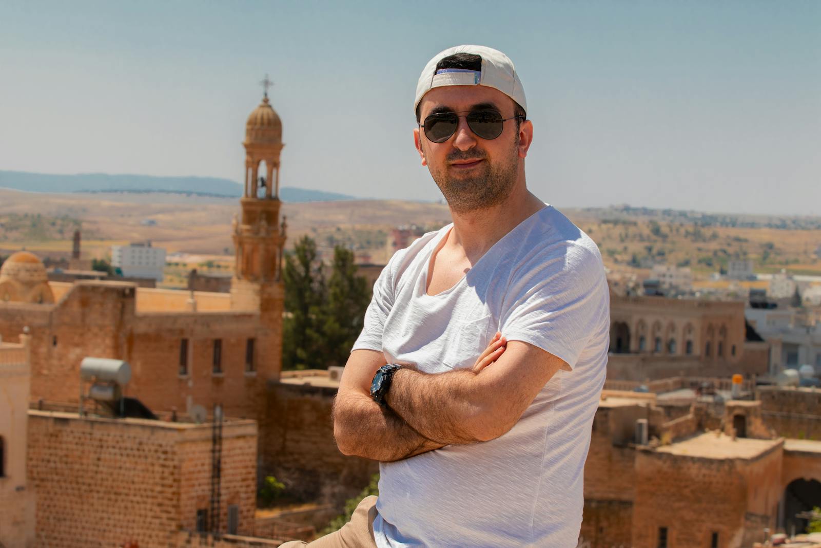 Man in white t-shirt and sunglasses posing near historic architecture on a sunny day.