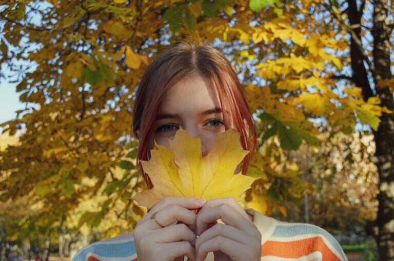 A young woman playfully holding yellow maple leaves against a backdrop of fall foliage.