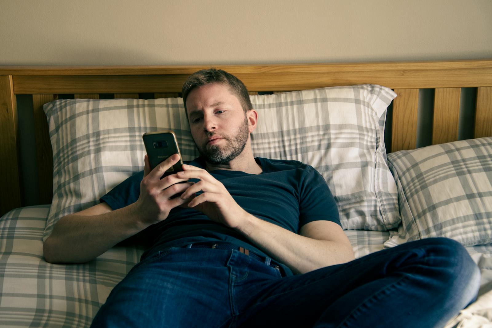 A man lying on a bed, focused on his smartphone, in a relaxed home setting.