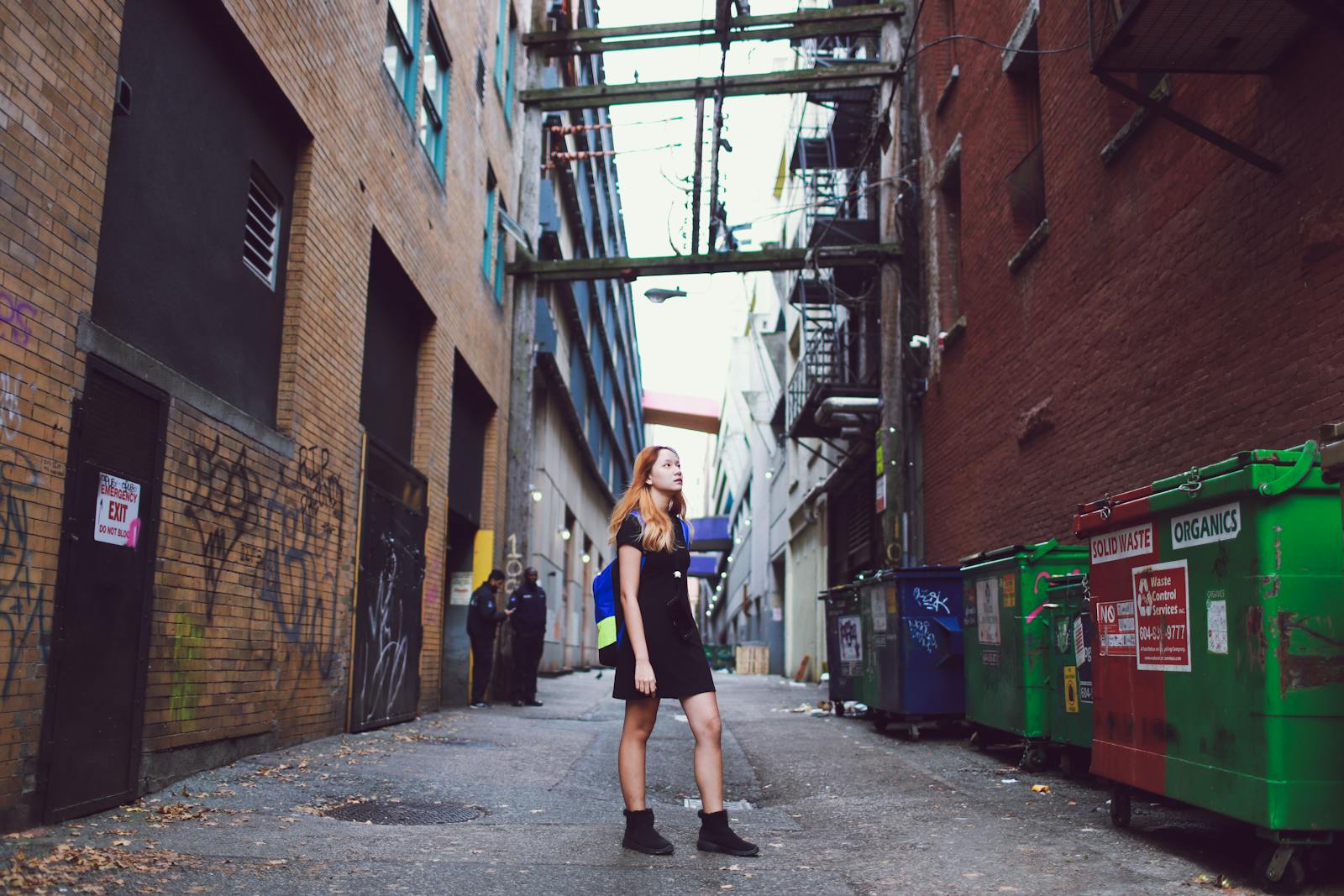 A woman stands in a graffiti-covered urban alley, surrounded by buildings and trash cans.