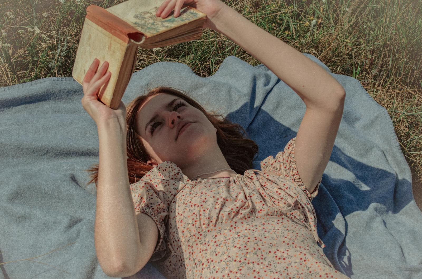 Young woman lying on a blanket reading a book outdoors in a grassy field.