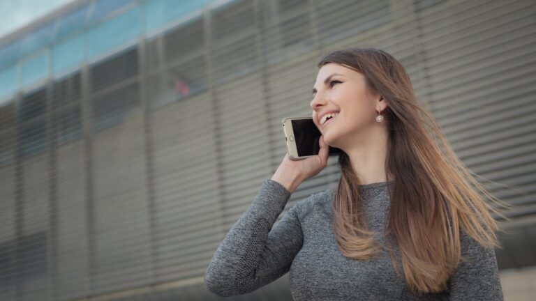 Woman in urban setting talking on phone, wearing casual attire, smiling.