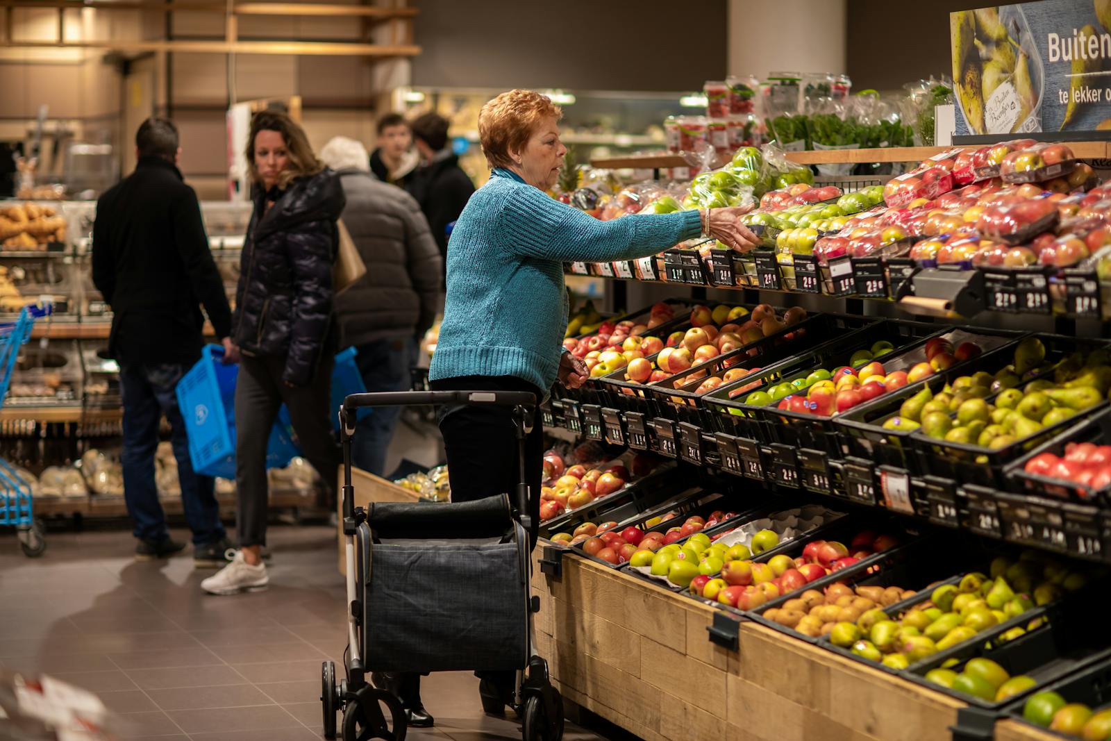 Elderly woman using walker, shopping for fresh produce in Utrecht supermarket.