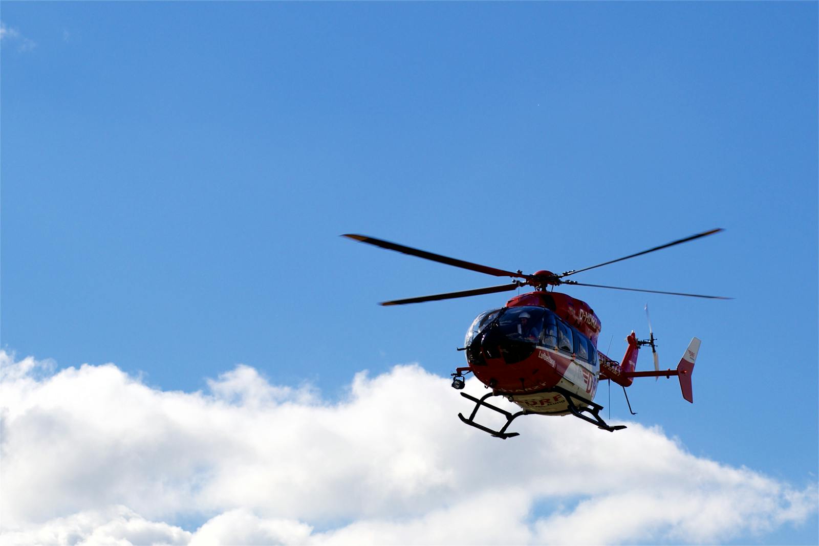 A red helicopter flies against a backdrop of clouds and clear blue sky.