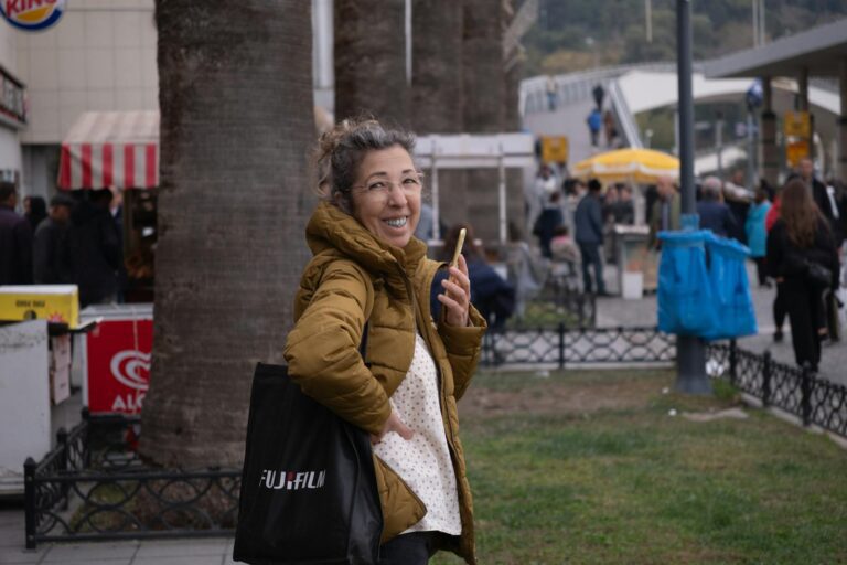 Smiling woman with phone and Fujifilm bag at bustling outdoor market in İzmir, Türkiye.