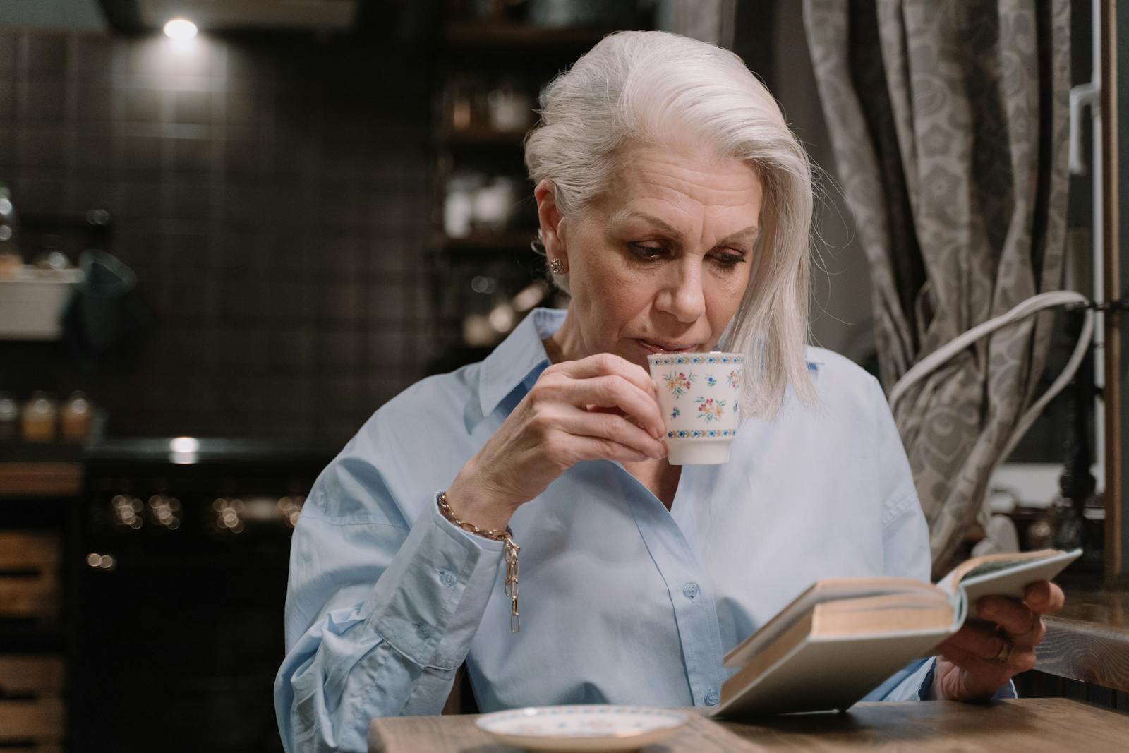 Elderly woman in a button-down shirt sipping tea while reading a book indoors.