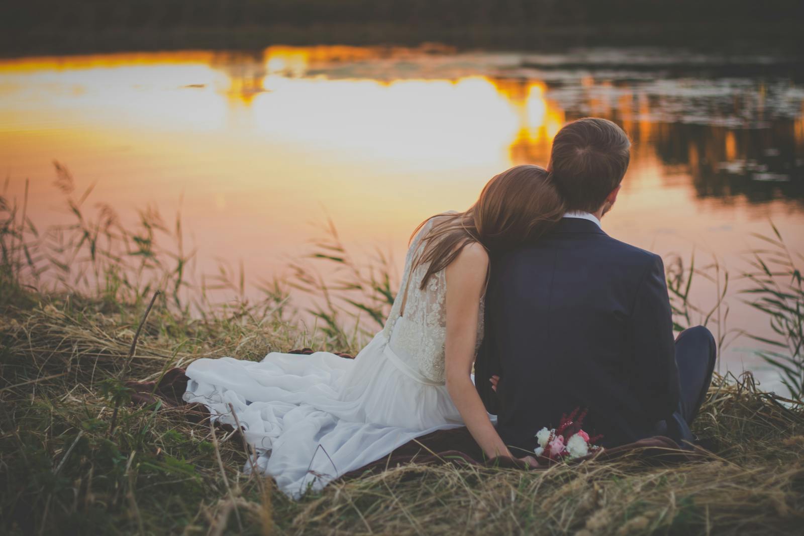 Newlywed couple sitting by a lake during sunset, embracing in a romantic setting.