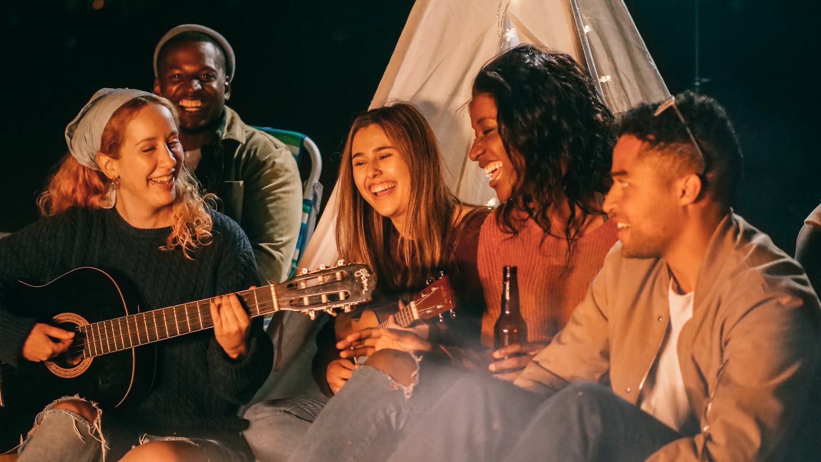 A diverse group of friends playing guitar and ukulele by a campfire at night, having fun and socializing.
