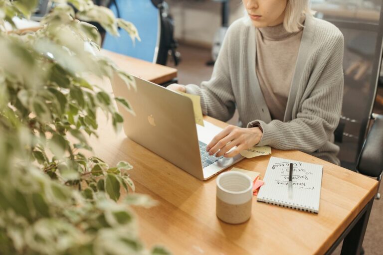 A woman working on a laptop at a wooden desk in a bright, modern office setting.